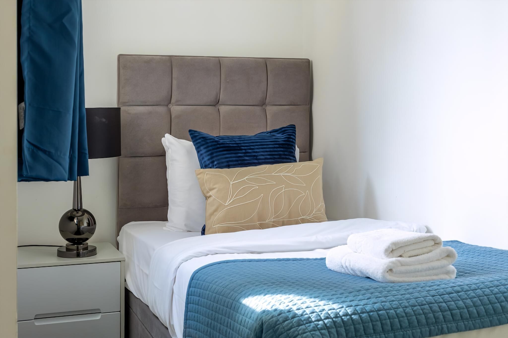 A neatly made bed with a blue quilt, a brown headboard, and a white pillow with a leaf pattern.