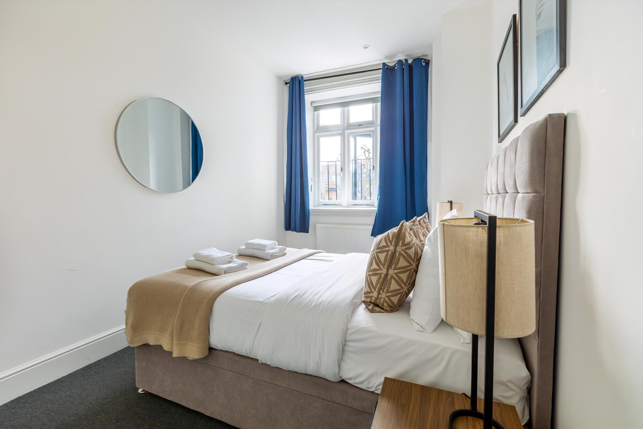 A neatly made bed with a beige blanket, a bedside lamp, and a round mirror on the wall.