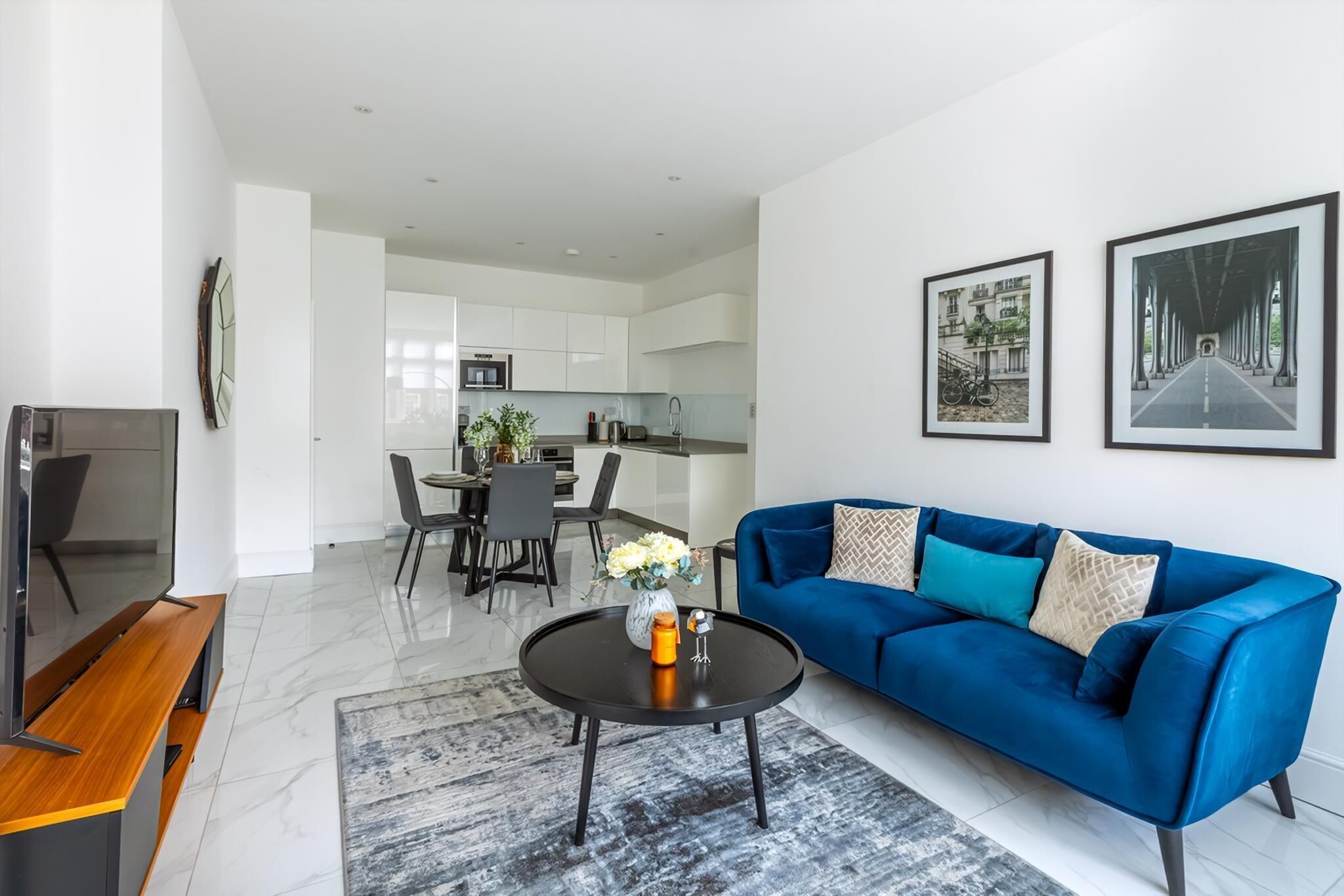 A modern living room with a blue sofa, a coffee table, and a TV. There is a dining area with chairs and a table, and a kitchen in the background.