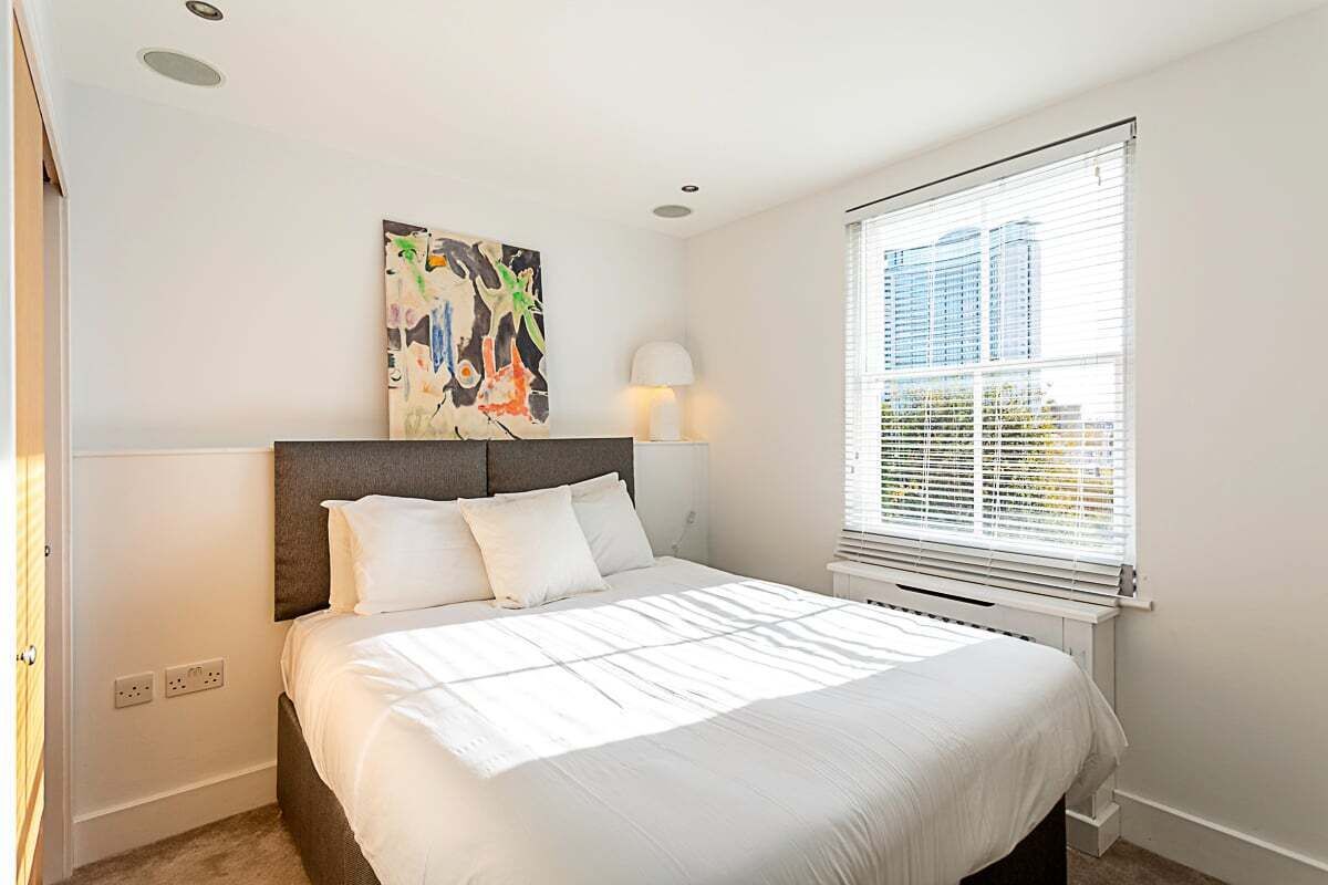 A bedroom with a large bed, a headboard, a window with blinds, a wall-mounted lamp, and a painting.