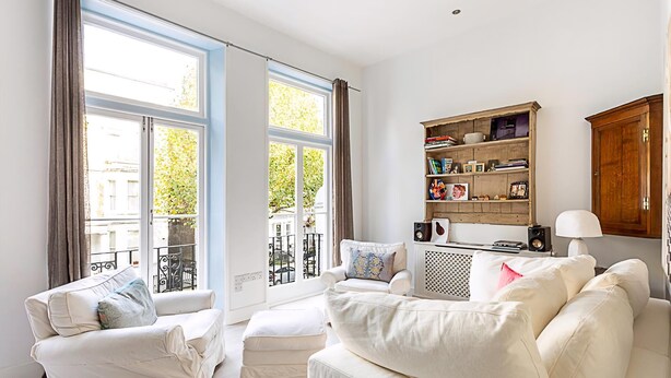 Een woonkamer met een bank, fauteuil en poef, een boekenkast, een lamp en een raam met gordijnen.