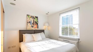 Een hotelkamer met een bed, een schilderij aan de muur, een raam met jaloezieën en een lamp.