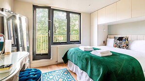Een moderne hotelkamer met een groot raam, een bed met een groene spreideken, een witte hoofdbord en een blauwe poef.