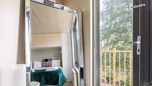 Een kleine slaapkamer met een bed, een tafeltje en een stoel. Een schuifdeur geeft toegang tot een buitenbalkon met een metalen hekwerk.