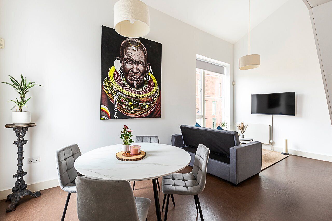 Un salon moderne avec une table à manger ronde, des chaises recouvertes d’un tissu gris, un canapé gris, un téléviseur à écran plat et un grand tableau abstrait accroché au mur.