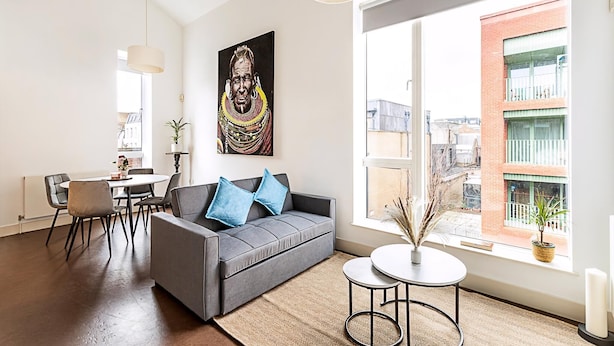 A modern living room with a grey sofa, a dining table, chairs, a large window with a view of buildings, and a painting on the wall. - Stylish Camden Flat Near Markets Tube (London)