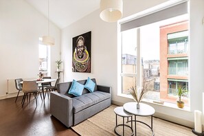 Un salon moderne avec un canapé gris, une table à manger, des chaises, une grande fenêtre donnant sur des immeubles et un tableau accroché au mur.