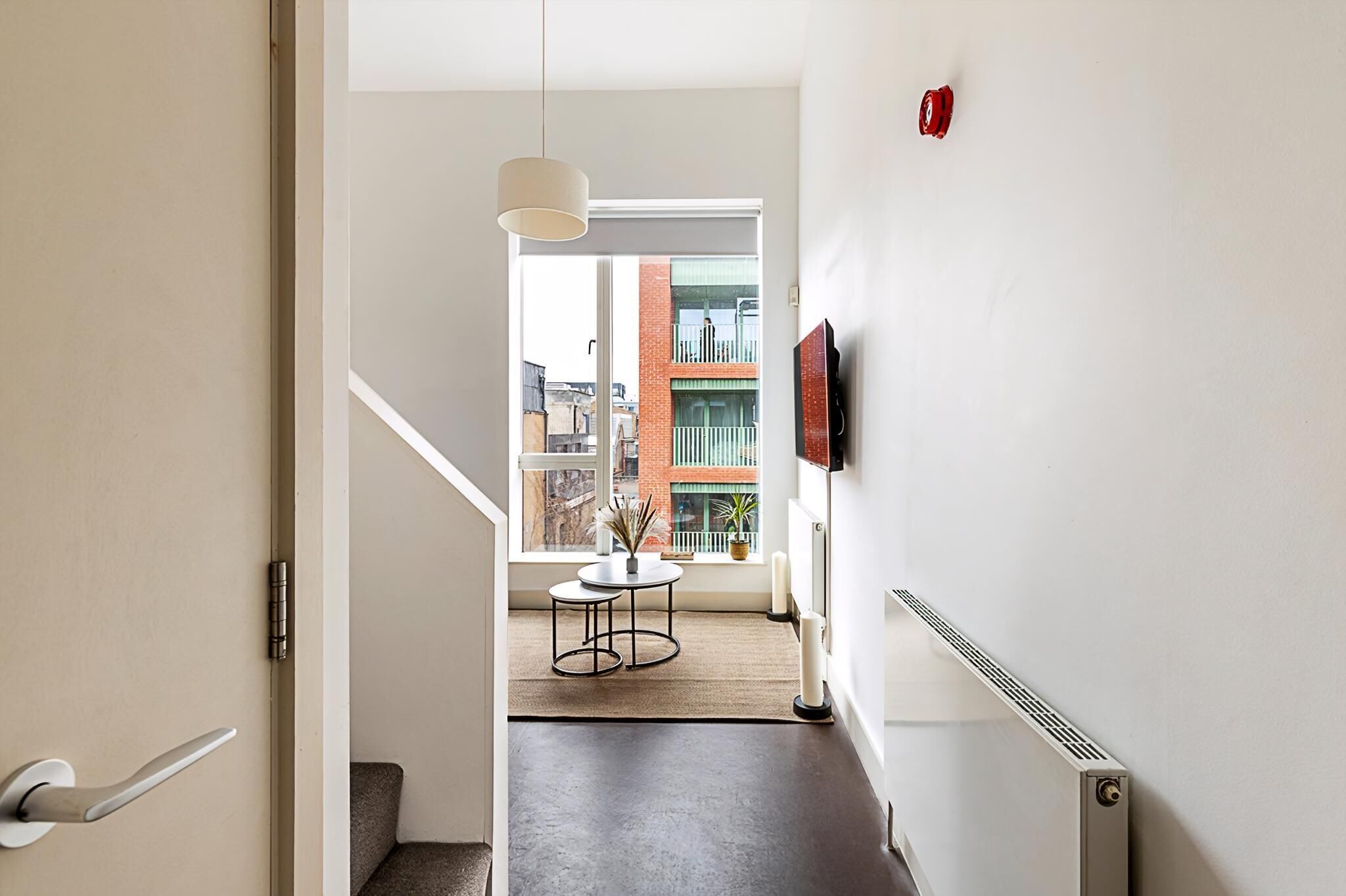 Un intérieur moderne avec un escalier, une petite table ornée d’une plante, un radiateur et une fenêtre donnant sur des immeubles.