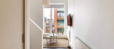 Un intérieur moderne avec un escalier, une petite table ornée d’une plante, un radiateur et une fenêtre donnant sur des immeubles.