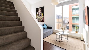 A modern apartment with a staircase, a gray sofa, a small round table, and a large window with a city view. - Stylish Camden Flat Near Markets Tube (London)