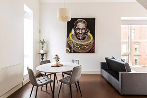A modern living room with a round dining table, grey upholstered chairs, a grey sofa, and a large painting on the wall.