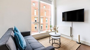 A modern living room with a sofa, a small round table, a flat-screen TV, and a large window with a view of a brick building. - Stylish Camden Flat Near Markets Tube (London)
