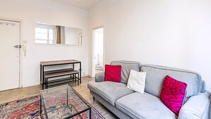 Un salon avec un canapé gris, des coussins rouges et blancs, une table basse en verre, un tapis à motifs, un banc et un miroir au mur.