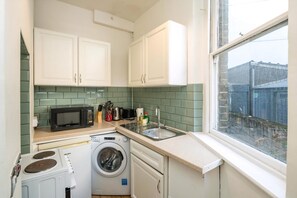 Een kleine keuken met witte kastjes, een magnetron, een wasmachine en een spoelbak.