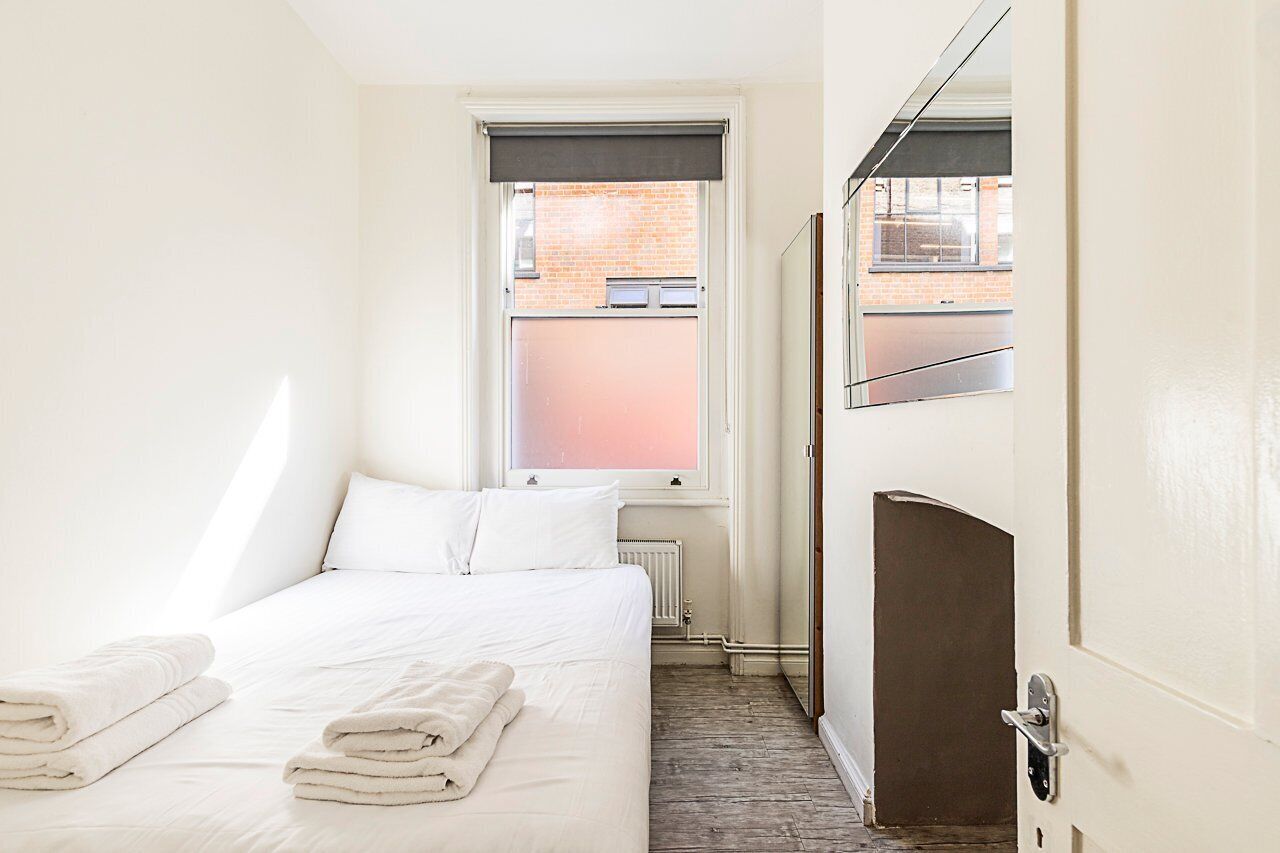 Une chambre d’hôtel avec un lit, un radiateur, une fenêtre donnant sur un immeuble en briques et une porte avec une poignée.