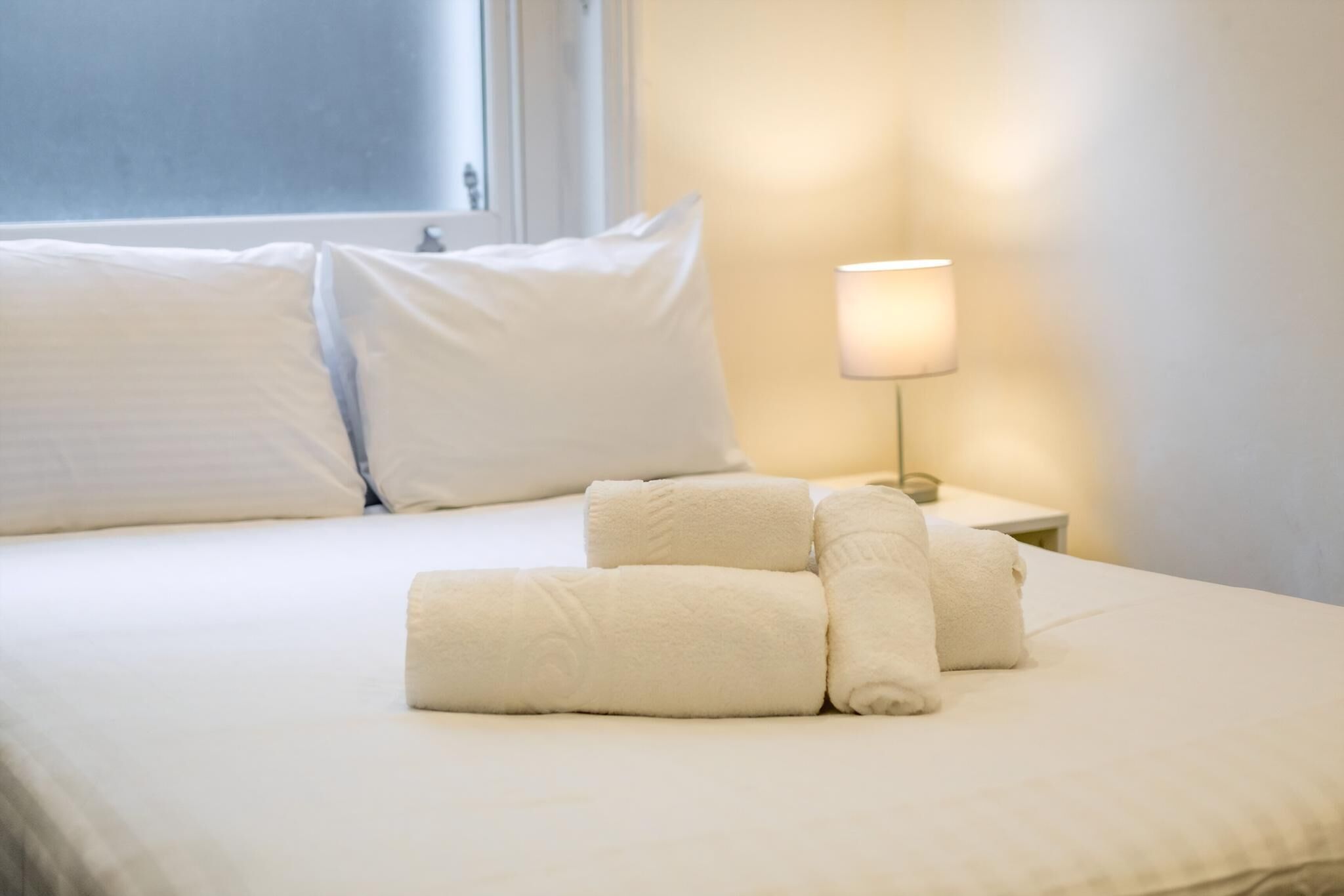Una cama bien hecha con sábanas y almohadas blancas, una lámpara de noche y una ventana con persianas.