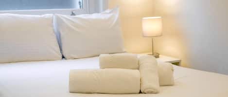 A neatly made bed with white linens and pillows, a bedside lamp, and a window with blinds.