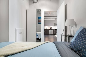 Un dormitorio con cama, mesita de noche, lámpara y puerta abierta que da a una cocina.