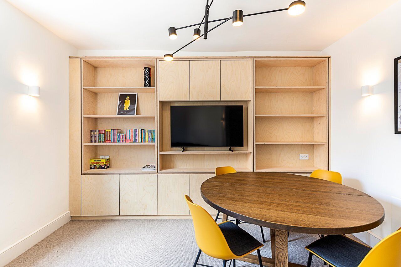 A modern living room with a wooden TV stand, a flat-screen TV, a round wooden dining table, and yellow chairs.