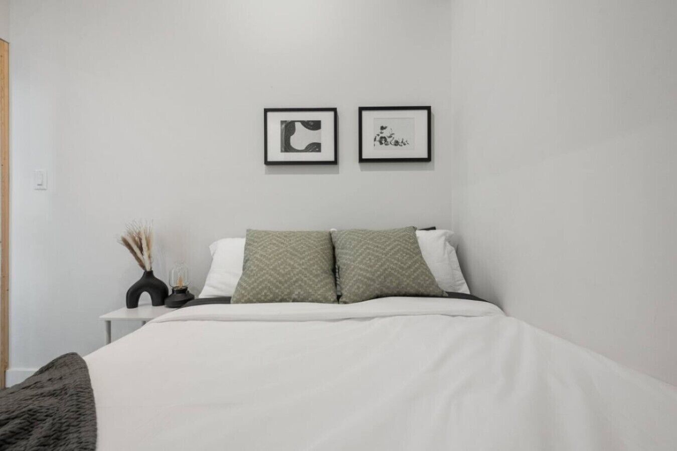 A bedroom with a bed, two framed pictures, a bedside table with a vase, and a lamp.