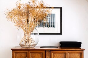 A clear glass vase with dried flowers on a wooden cabinet, a framed photograph on the wall, and a black object on the cabinet.