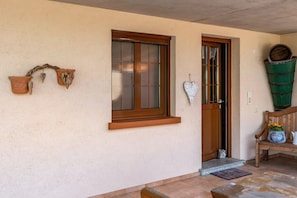 A house entrance with a wooden door, a window with a wooden frame, a bench, and a potted plant.