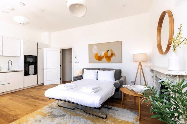 A modern hotel room with a bed, bedside table, lamp, and a view of the kitchen.