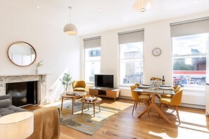 Uma sala de estar moderna com lareira, televisão, área de jantar com cadeiras amarelas e grandes janelas com vista para a cidade.
