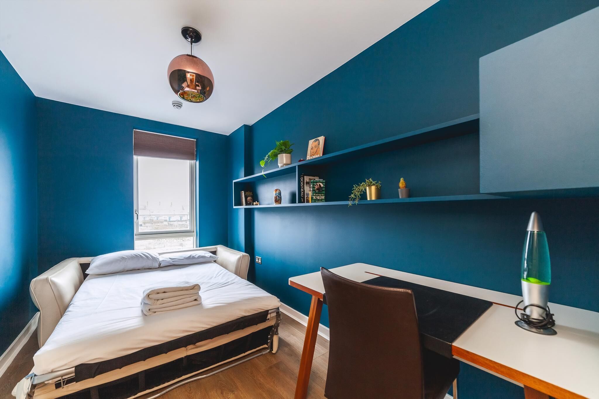 A room with a bed, a desk, a chair, a lava lamp, and shelves with plants and books.