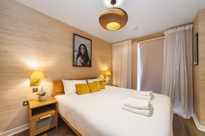 A hotel room with a large bed, two bedside lamps, a painting on the wall, and a window with curtains. - Cosy 2-bed Poplar Flat w/ City Views (London)