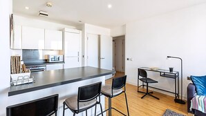 Ett modernt kök med vita skåp, en köksö med barstolar, en matplats med ett skrivbord och en stol samt en liten sittgrupp med en soffa.