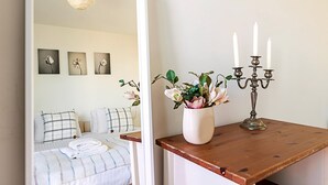 Ett sovrum med en säng, kuddar, ett sängbord med en vas med blommor och en ljusstake.