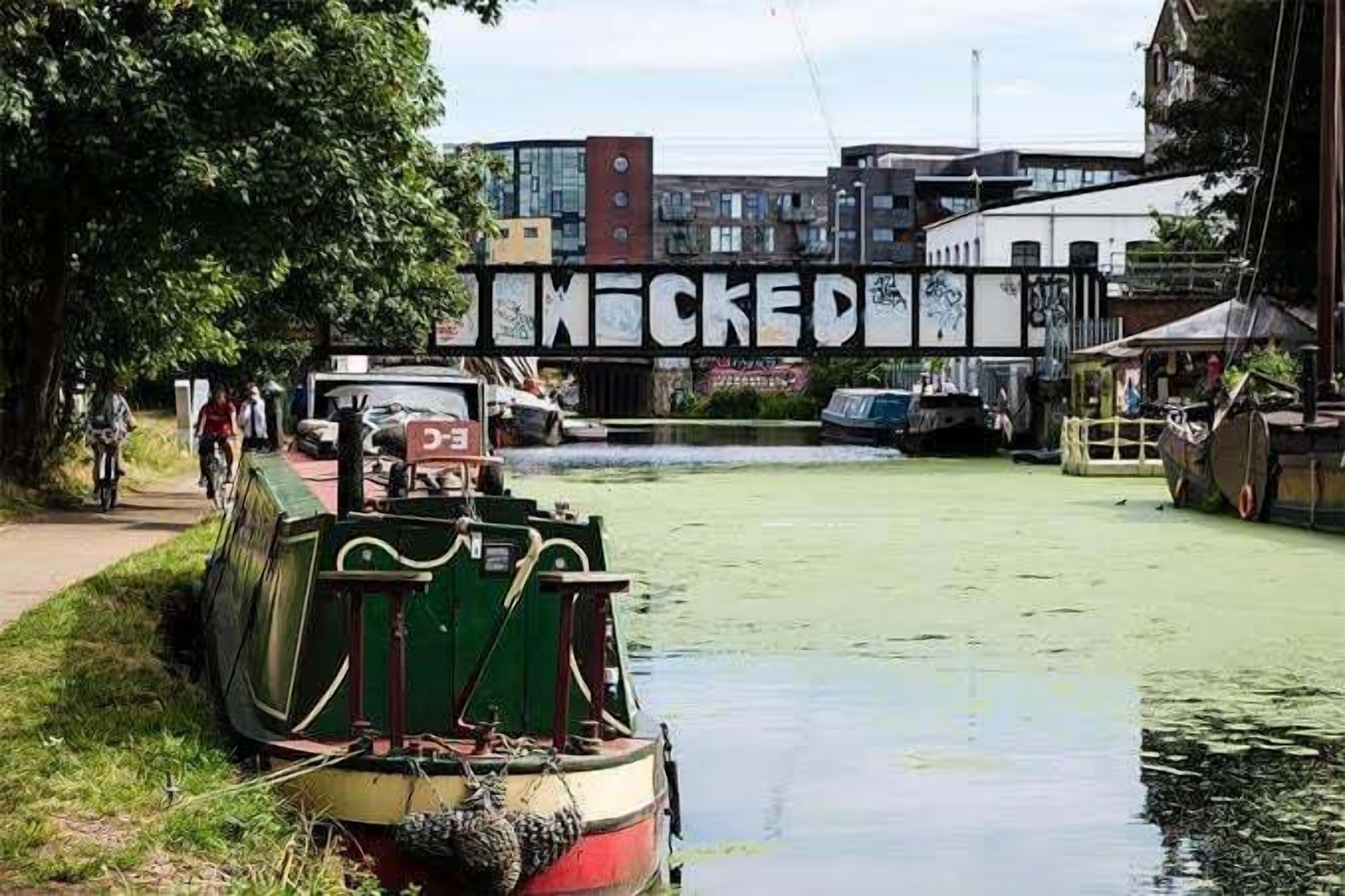 Ein schmaler Kanal mit einem grünen Boot, das am linken Ufer liegt, und einer Brücke mit Graffiti im Hintergrund.