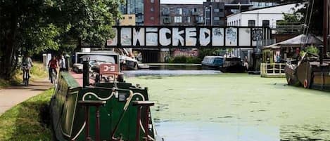 A narrow canal with a green boat docked on the left and a bridge with graffiti in the background.