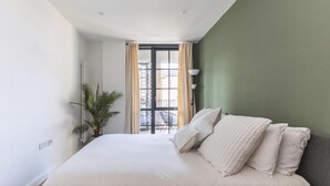 A bedroom with a bed, a potted plant, a radiator, a window with curtains, and a view of buildings outside. - Stylish Hackney Wick Apartment With Terrace (London)