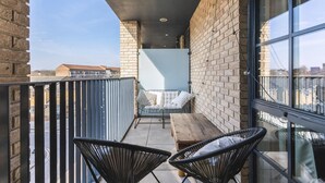 A balcony with a wooden table and two black chairs, a cushioned bench, and a view of the city through large windows. - Stylish Hackney Wick Apartment With Terrace (London)