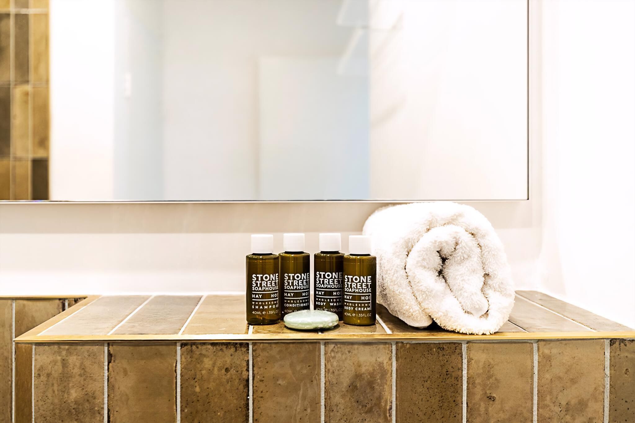 A bathroom with a tiled sink, four bottles of Stone Street Soaphouse products, and a rolled towel.