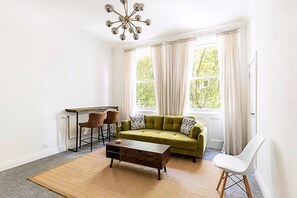 A living room with a green sofa, a wooden coffee table, a white chair, and a bar stool. - Elegant Knightsbridge Flat Near Harrods (London)
