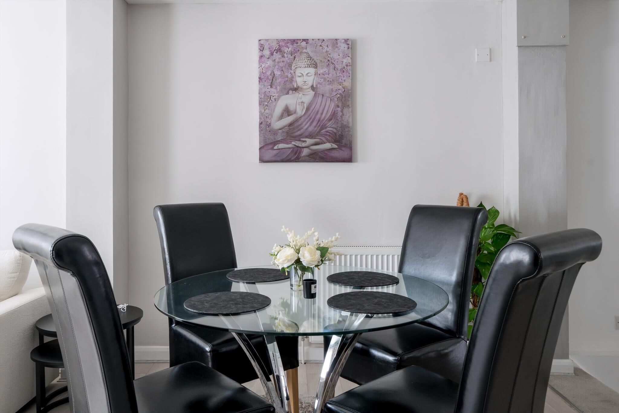 A dining area with a glass table surrounded by black leather chairs, a vase with white flowers, and a Buddha painting on the wall.