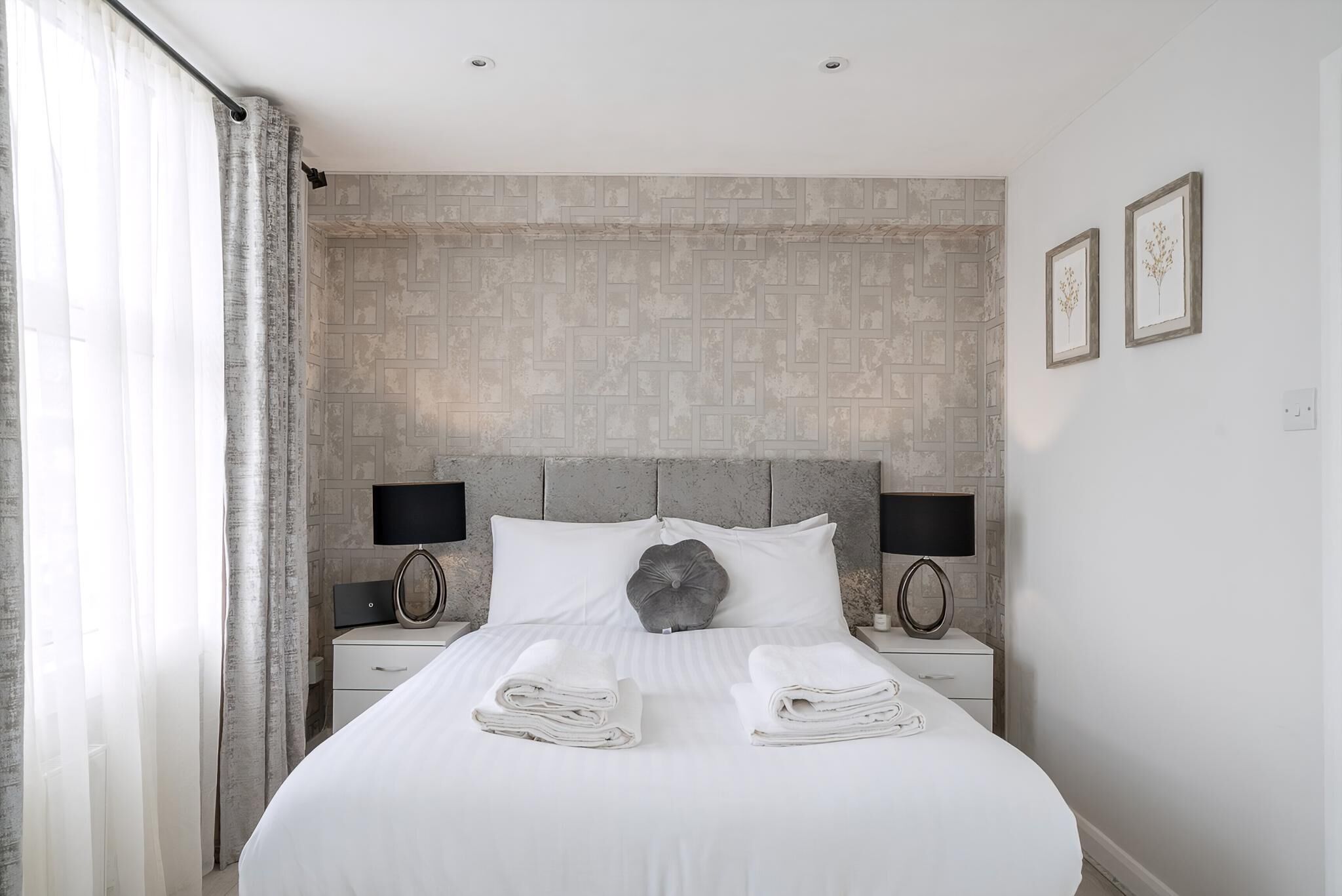 A neatly made bed with folded towels, a grey headboard, two bedside lamps, and a wall with a geometric pattern.