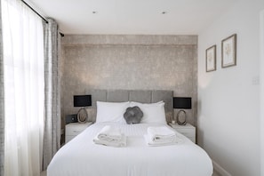 A neatly made bed with folded towels, a grey headboard, two bedside lamps, and a wall with a geometric pattern. - Modern South Wimbledon Stay (London)