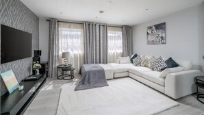 A modern living room with a white sofa, a flat-screen TV, a coffee table, and a large window with curtains. - Modern South Wimbledon Stay (London)