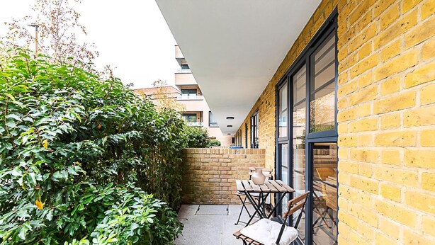 An outdoor balcony with a brick wall, a hedge, and two wooden tables with chairs. - Cosy and Stylish Studio in the Heart of Greenwich (London)