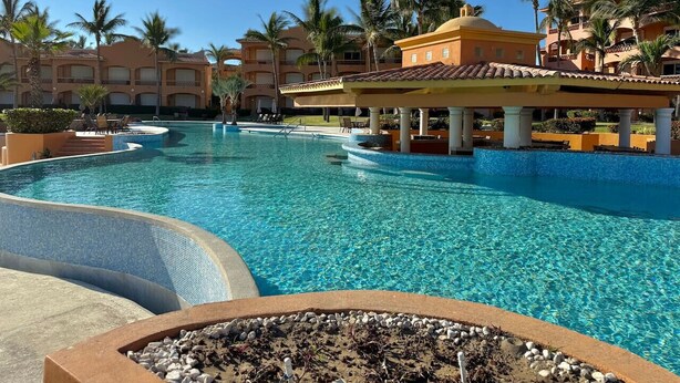 A resort pool with a clear blue water, surrounded by palm trees and resort buildings. - Depa in EDM first floor beachfront By GPS (Mazatlán)