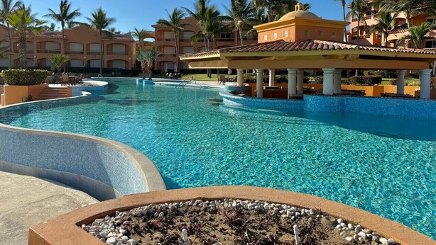 Un'ampia piscina cristallina con zona a vasca profonda, circondata da palme e edifici di un hotel con balconi.