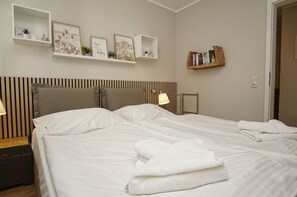 En pænt redt seng med hvide sengetøj, et stribet sengegavl og et natbord med en lampe. Der er vægmonterede hylder med dekorative genstande og en lille bogreol.