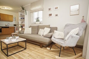 Uma sala de estar com sofá, poltrona de balanço, mesa de centro, estante de livros e área de cozinha.