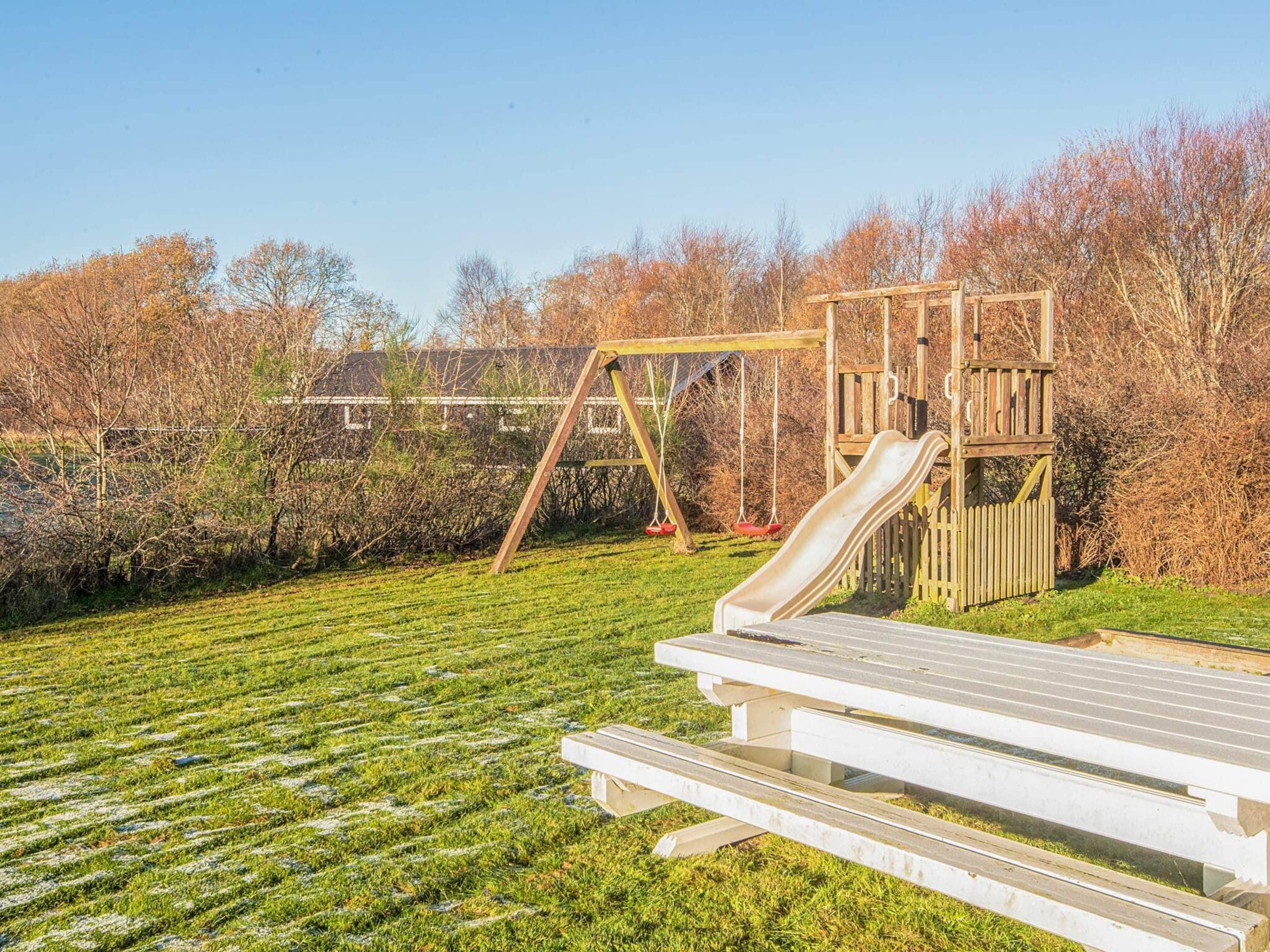 Un parque infantil con tobogán y columpios, rodeado de césped y árboles.