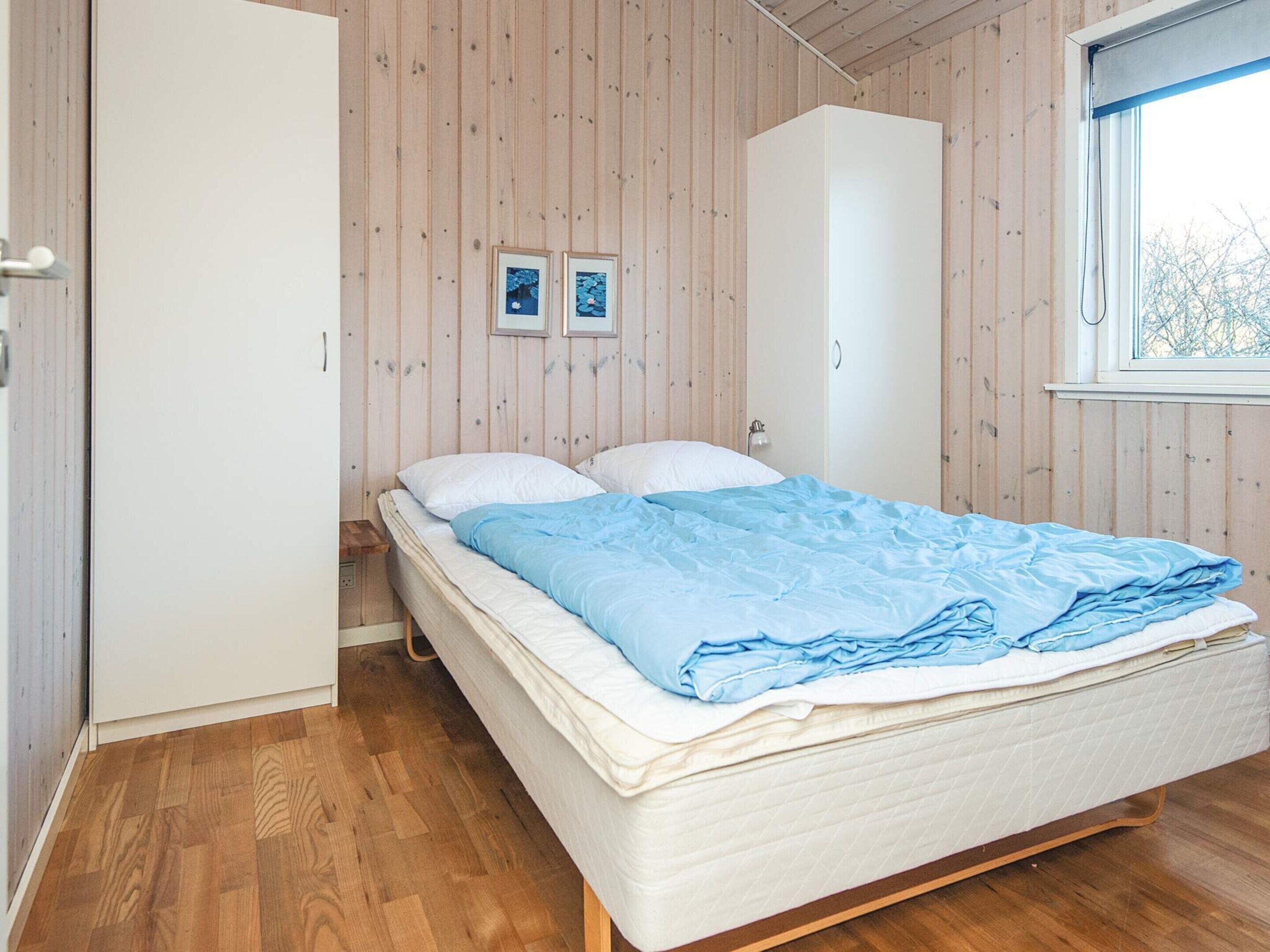 A bedroom with a wooden floor, a bed with a blue blanket, two framed pictures on the wall, and a window with a view of trees.