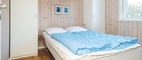 A bedroom with a wooden floor, a bed with a blue blanket, two framed pictures on the wall, and a window with a view of trees.